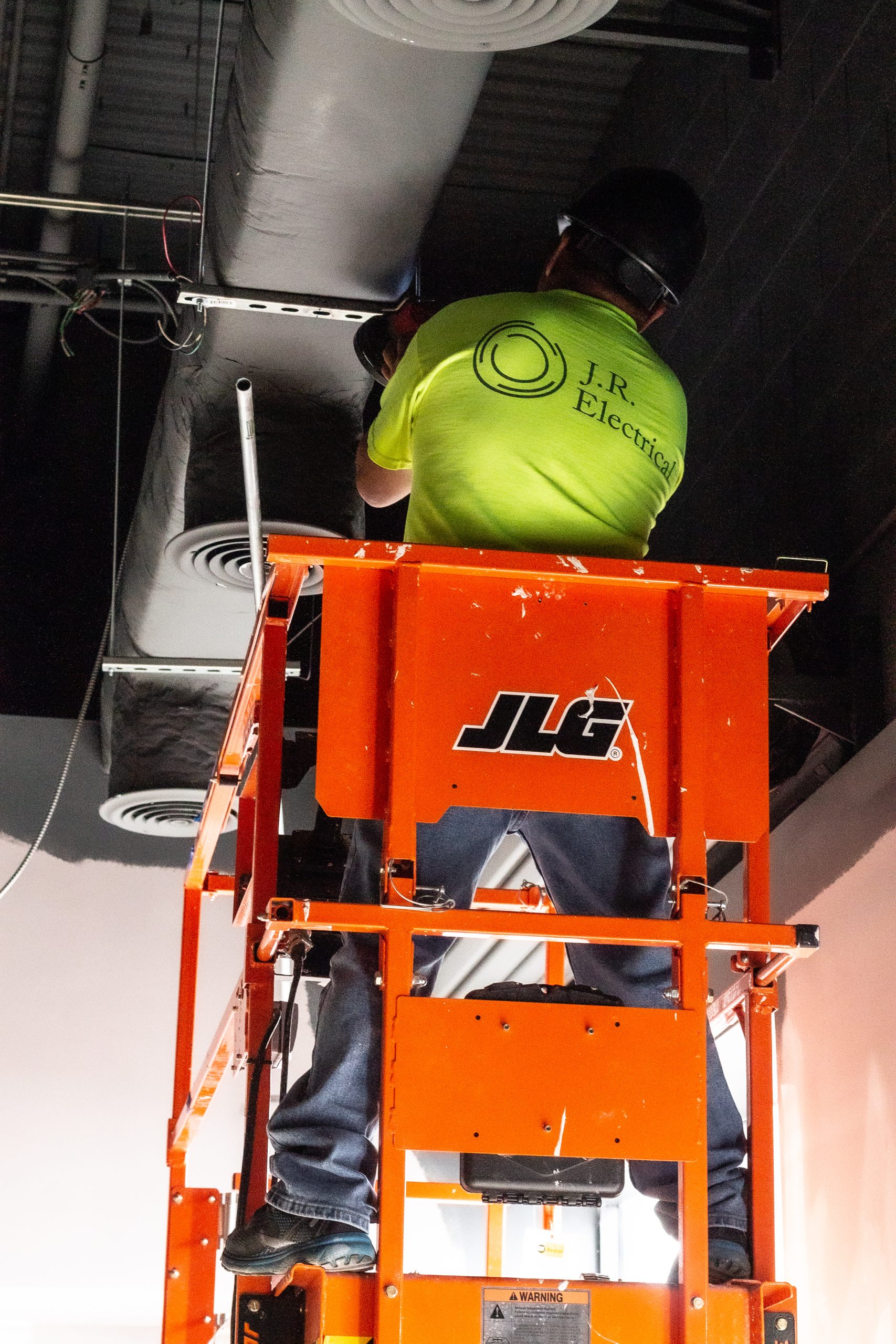 An electrician wearing a bright yellow shirt with the logo 'J.R. Electrical' works on ceiling ducts from an orange JLG lift. The worker is focused on adjusting metal brackets and wires, emphasizing the maintenance and infrastructure of the building. The ceiling features large ventilation ducts and wiring, indicating an industrial or commercial setting.